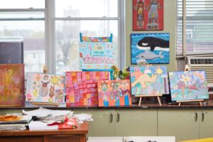 A photograph of various paintings made by BEING artists on easels at the Studio.