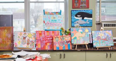 A photograph of various paintings made by BEING artists on easels at the Studio.