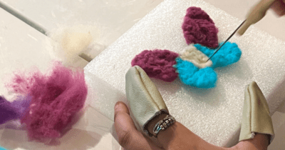 A close-up photo of someone felting a blue purple butterfly on a white sponge.